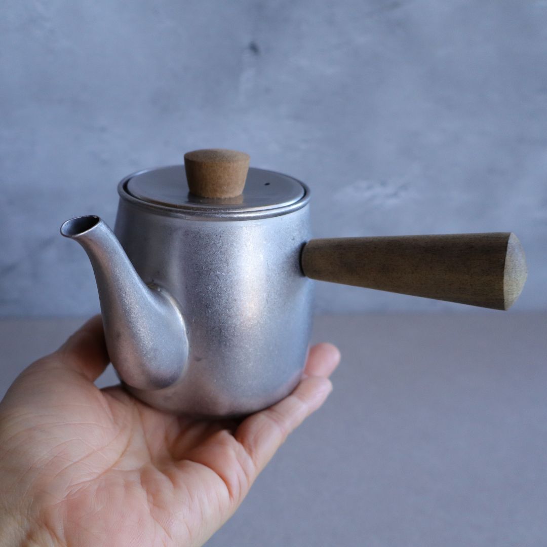 A person holding the compact stainless steel teapot with a wooden side handle, highlighting its small size