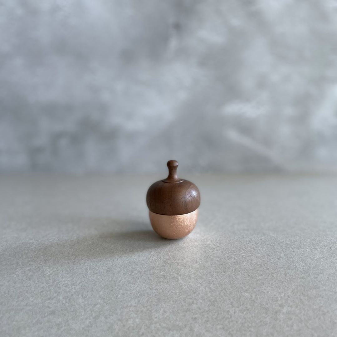 A small acorn-shaped healing bell with a rose gold base and a wooden top, placed on a smooth surface against a soft, neutral background.