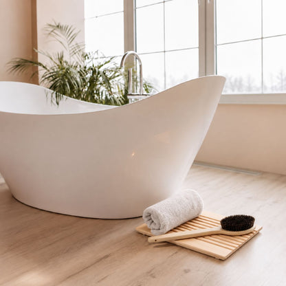 A modern freestanding bathtub in a bright bathroom setting with a wooden floor, large window, green plant, and a rolled towel with a wooden brush on a slatted mat.