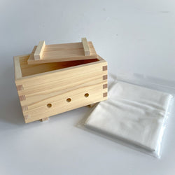 A wooden Hinoki tofu mold with an accompanying tofu cloth placed on a white background. The tofu mold features a simple design made from wooden slats, with three ventilation holes at the bottom. The tofu cloth is neatly packaged in a clear plastic bag.