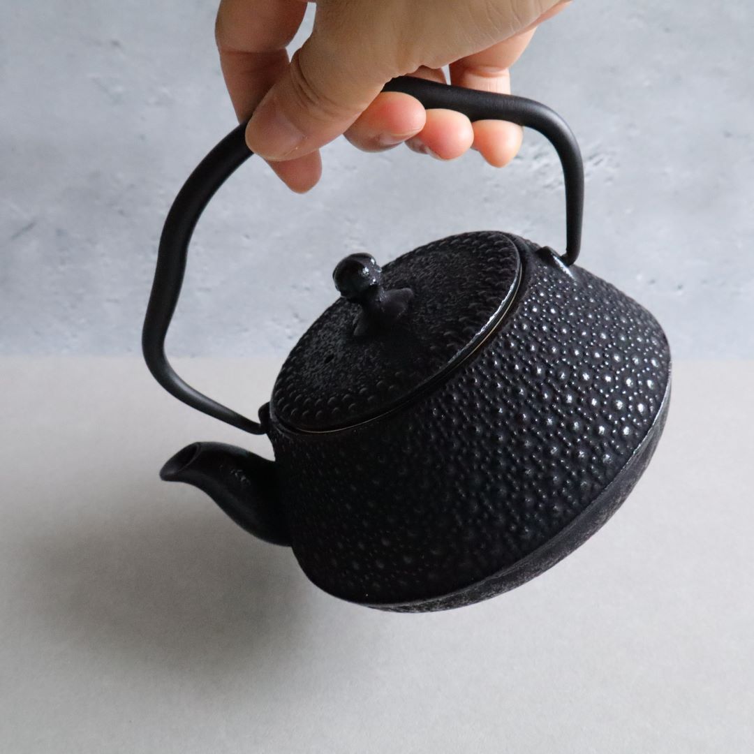 A hand holding the handle of a Nambu Tekki teapot, demonstrating its ergonomic design and traditional hexagonal pattern. The teapot is set against a neutral background.