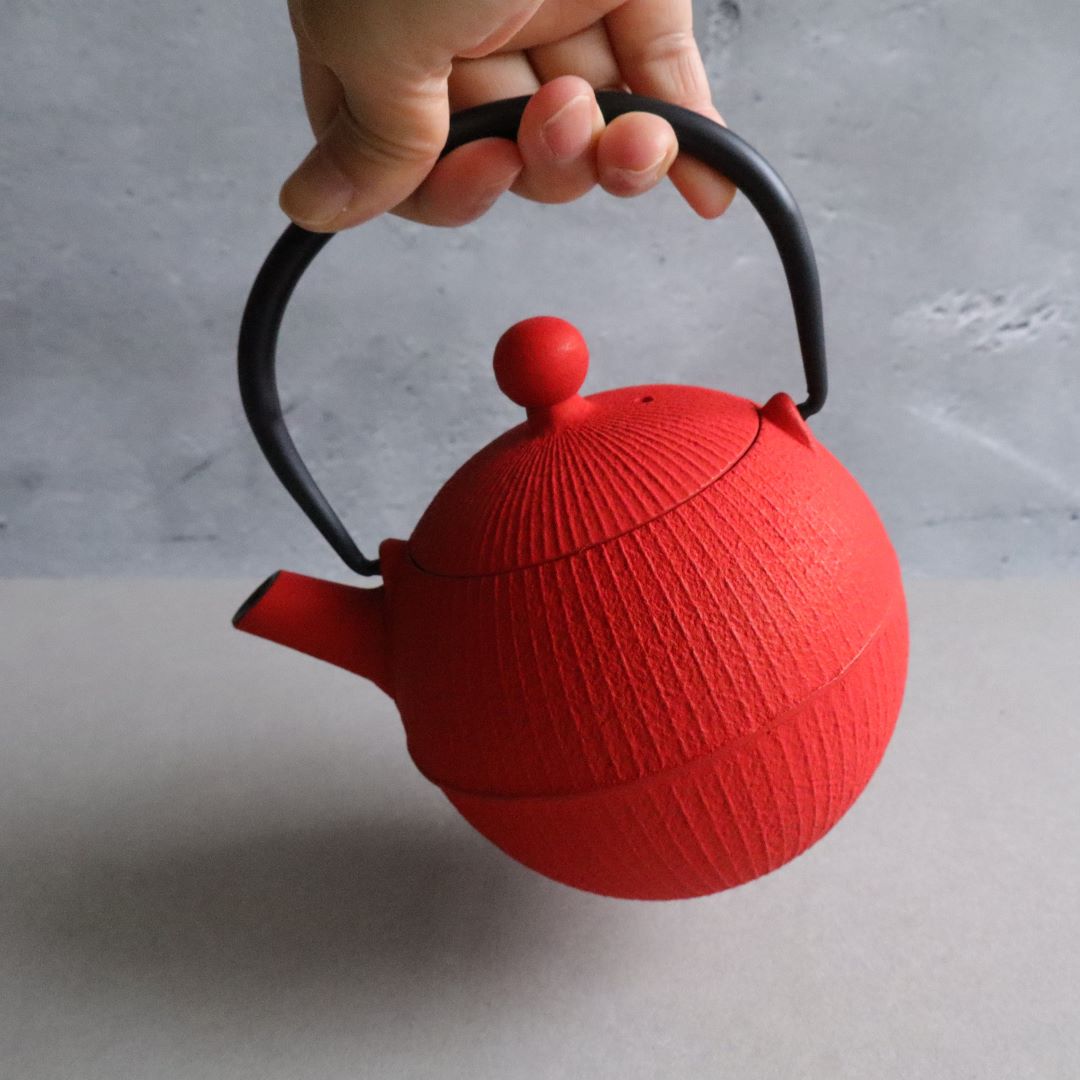 A hand holding the handle of a vibrant red Nambu Tekki teapot, shaped like a traditional Japanese temari ball. The teapot features a black handle and a unique design.