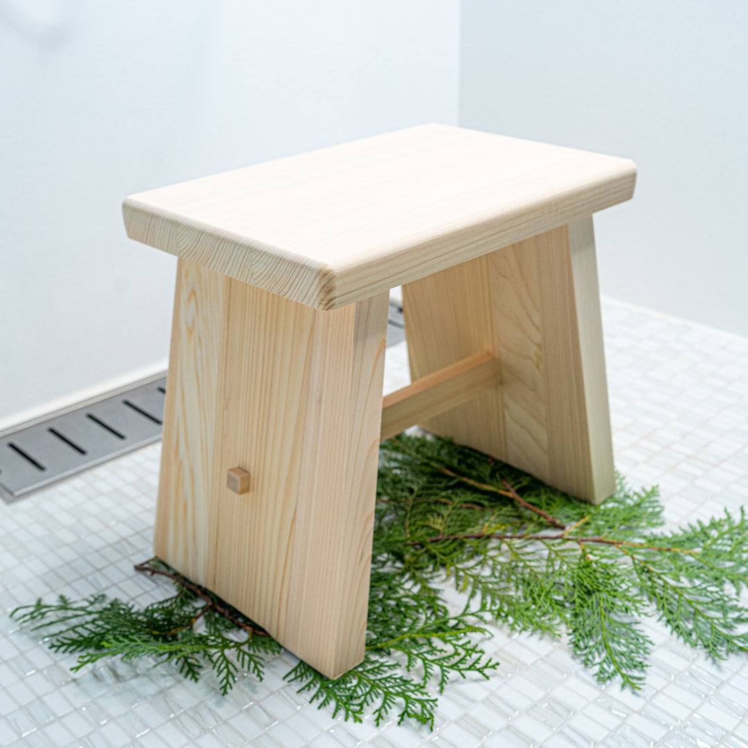 A beautifully crafted Japanese bath stool made from high-quality cypress wood. The stool features a smooth, pale wood finish with rounded edges, providing both elegance and functionality. It is set against a white tiled background with green foliage underneath, highlighting its natural and eco-friendly design."