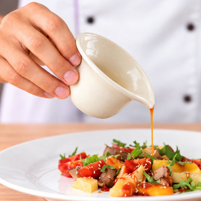 A close-up of a hand pouring a glossy light beige sauce from a small ceramic pouring cup onto a plated salad with vegetables, sesame seeds, and pieces of meat, set against a blurred chef&