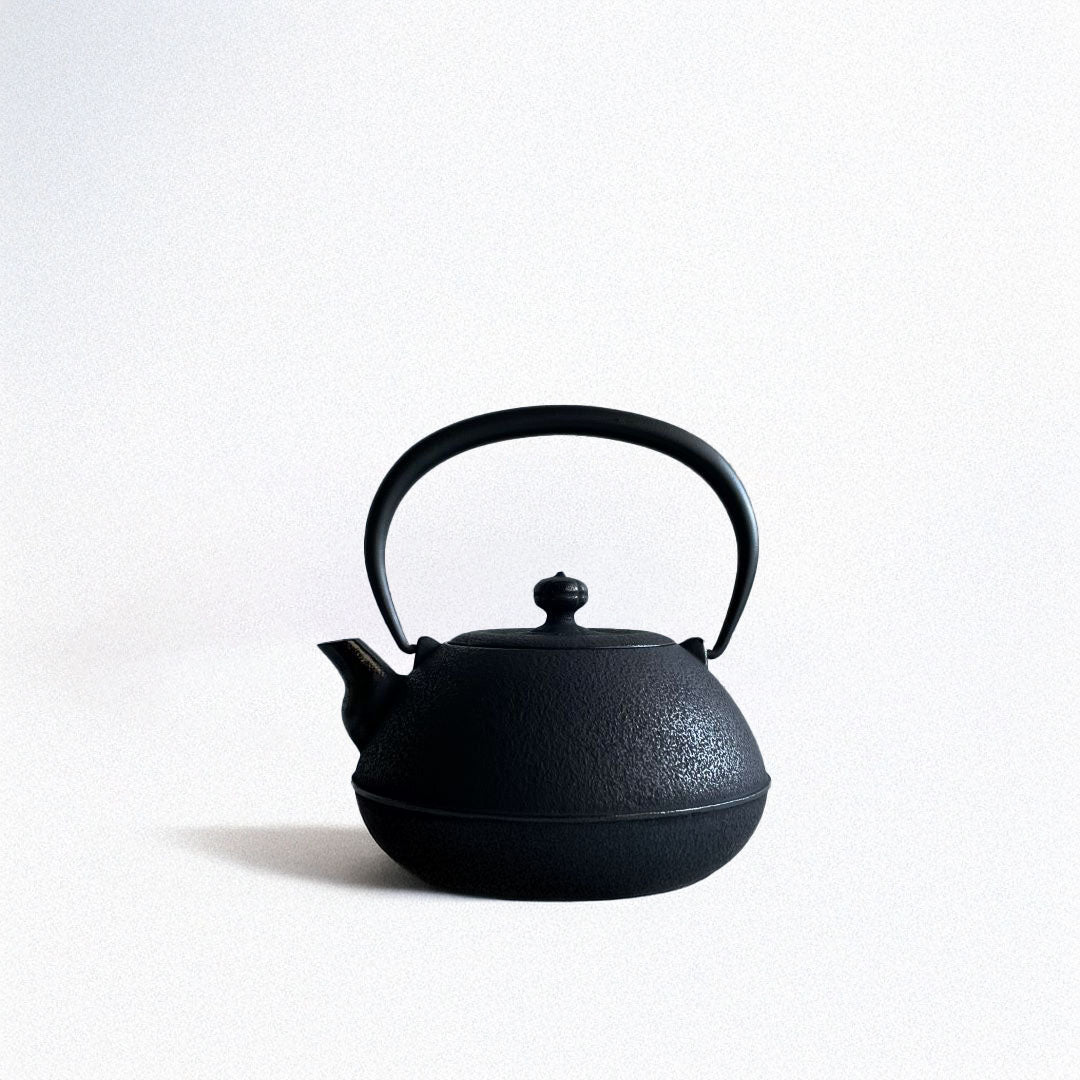 This is a black cast iron Tetsubin kettle with a lightly rough surface on the body. The iron handle has a smooth surface and is standing upright. The kettle is placed in the center of a space with grey walls and a grey surface, seen from a front view.