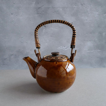 Handmade Japanese ceramic teapot with a warm brown glaze and a woven bamboo handle. The teapot features a classic Kyusu-style design, perfect for brewing loose-leaf tea. Its glossy finish and traditional craftsmanship make it both functional and decorative.