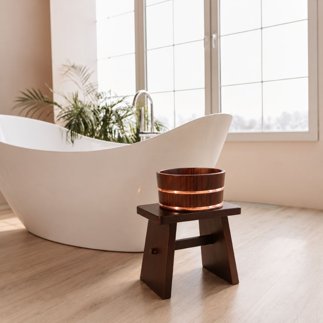 A dark brown wooden bath bowl with a glowing warm light inside, displayed on a small wooden stand in a modern bathroom setting with a white freestanding bathtub and large windows.