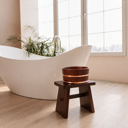 A dark brown wooden bath bowl with a glowing warm light inside, displayed on a small wooden stand in a modern bathroom setting with a white freestanding bathtub and large windows.