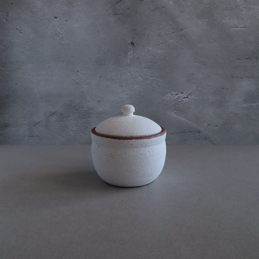 A white handmade Japanese Shigaraki ceramic salt cellar with a rustic brown-edged lid, centered on a grey textured background.