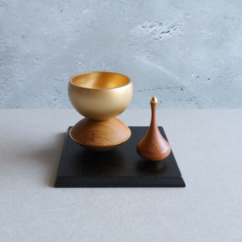 A Japanese Buddhist singing bowl set, featuring a golden bowl on a wooden base with a wooden striker, displayed on a black platform against a light gray background.