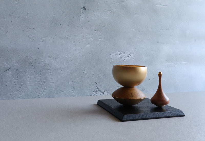 Side view of a Japanese Buddhist singing bowl set, showcasing a golden bowl resting on a wooden base next to a wooden striker, all placed on a black platform against a light gray background.