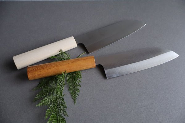 Two Japanese Santoku knives, one with a light wooden handle and the other with a dark brown wooden handle, displayed side by side on a grey surface with green foliage.