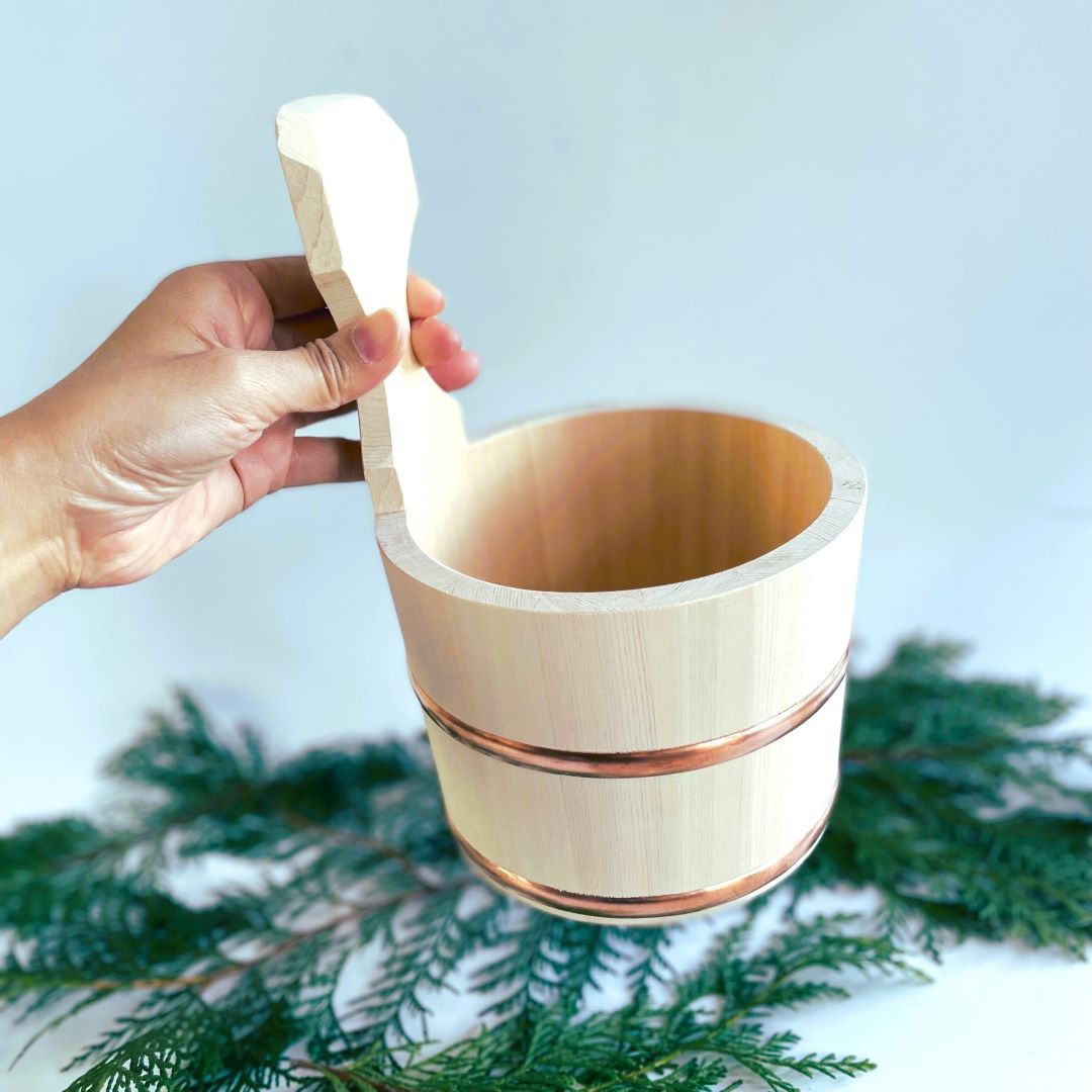 A hand holding a traditional Japanese wooden bath bucket with a handle. The bucket is made from light-colored wood, featuring two metal bands around its body for added strength. It is set against a backdrop of green foliage, highlighting its natural and eco-friendly design.