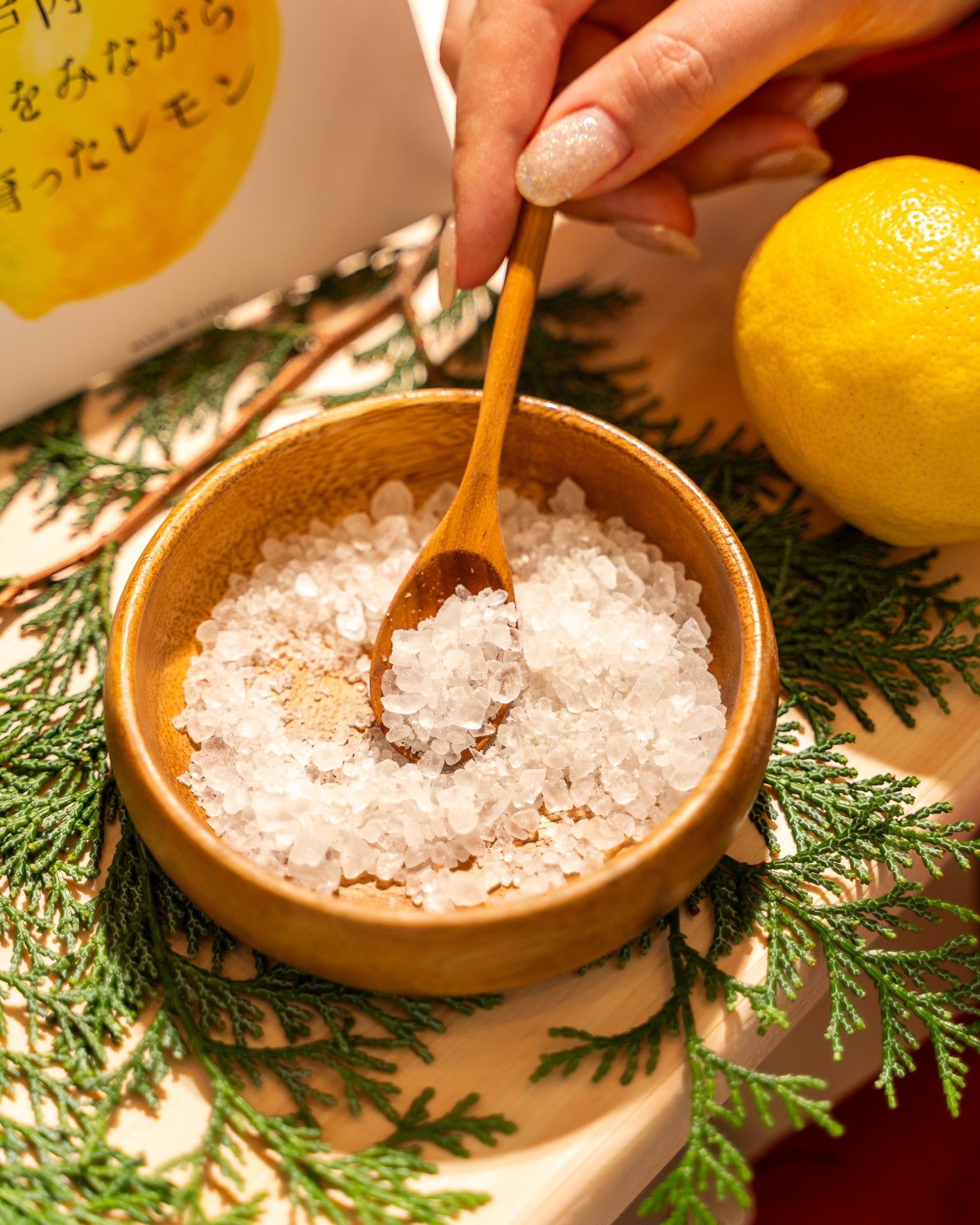 Wooden bowl with sea salt, wooden spoon, lemon, and greenery on a textured surface.