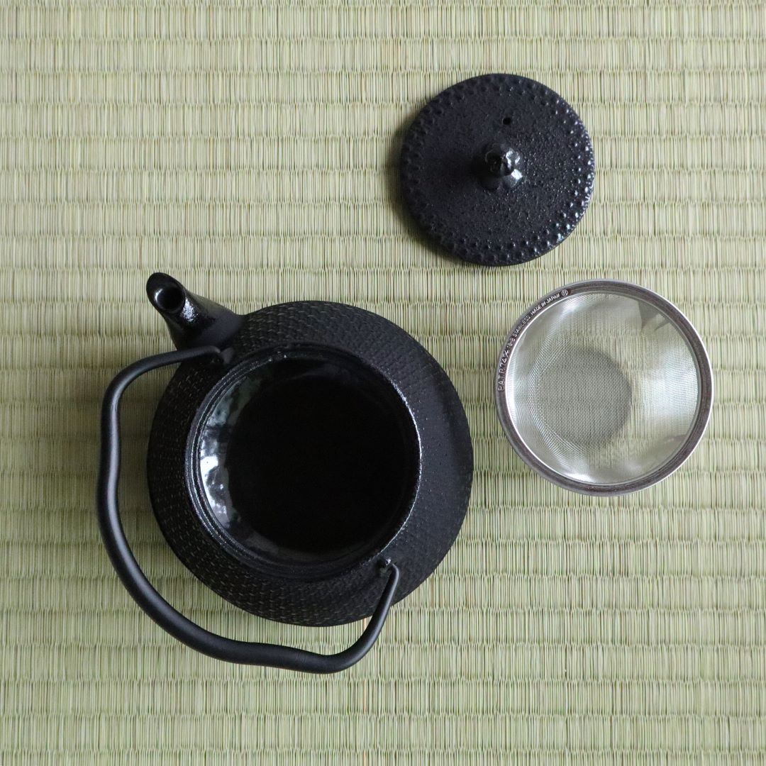 Top view of a Nambu Tekki teapot with the lid removed, showcasing the interior and the stainless steel mesh strainer. The teapot, lid, and strainer are arranged on a traditional tatami mat background, highlighting the intricate hexagonal pattern and craftsmanship of the teapot.