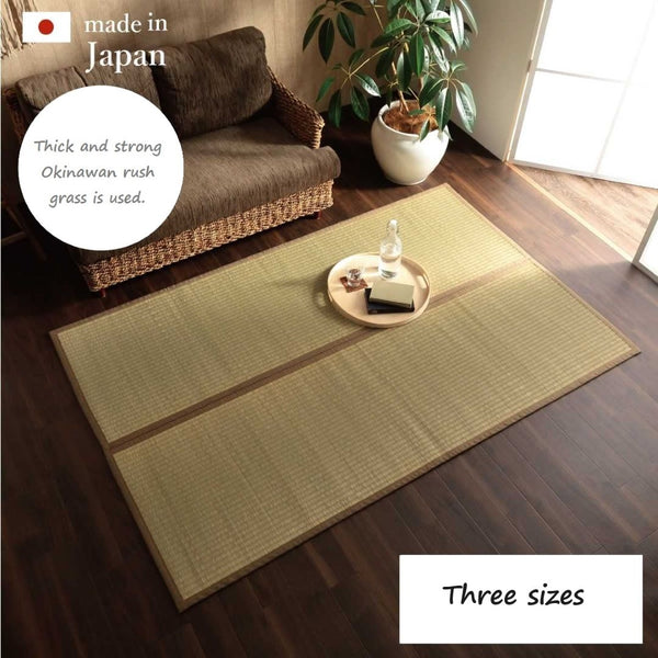 A cozy living room with a wooden floor featuring a rectangular tatami mat made from thick and strong Okinawan rush grass. The mat is placed in front of a wicker sofa with brown cushions, and there is a tray with a book, glass bottle, and cup on the mat. A large potted plant is positioned near a window, allowing natural light to brighten the room. The text "made in Japan" and "Three sizes" is displayed, emphasizing the product's origin and size options.