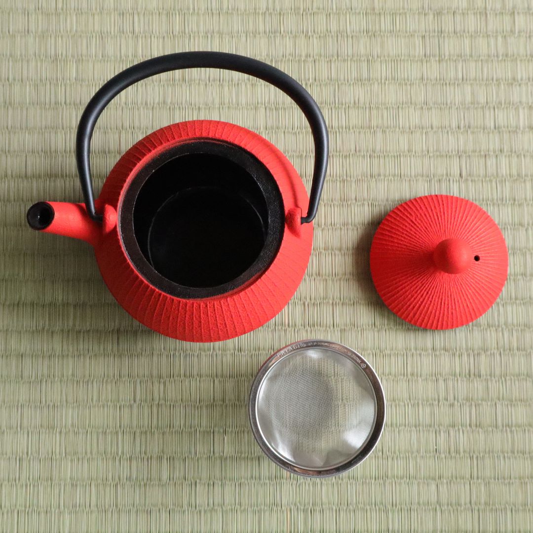 Top view of a red Nambu Tekki teapot, showcasing the enamel-coated interior and the stainless steel mesh strainer. The teapot, lid, and strainer are arranged on a traditional tatami mat background.