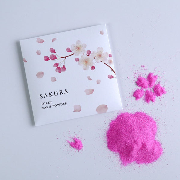 Sakura Milky Bath Powder package displayed alongside cherry blossom-shaped pink bath powder on a white surface.