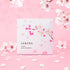A beautifully packaged Sakura Milky Bath Powder with a delicate cherry blossom illustration on the front. The package is set against a pink background with scattered sakura petals, creating a soft and elegant Japanese aesthetic. Perfect for a luxurious bath experience infused with the gentle fragrance of cherry blossoms.