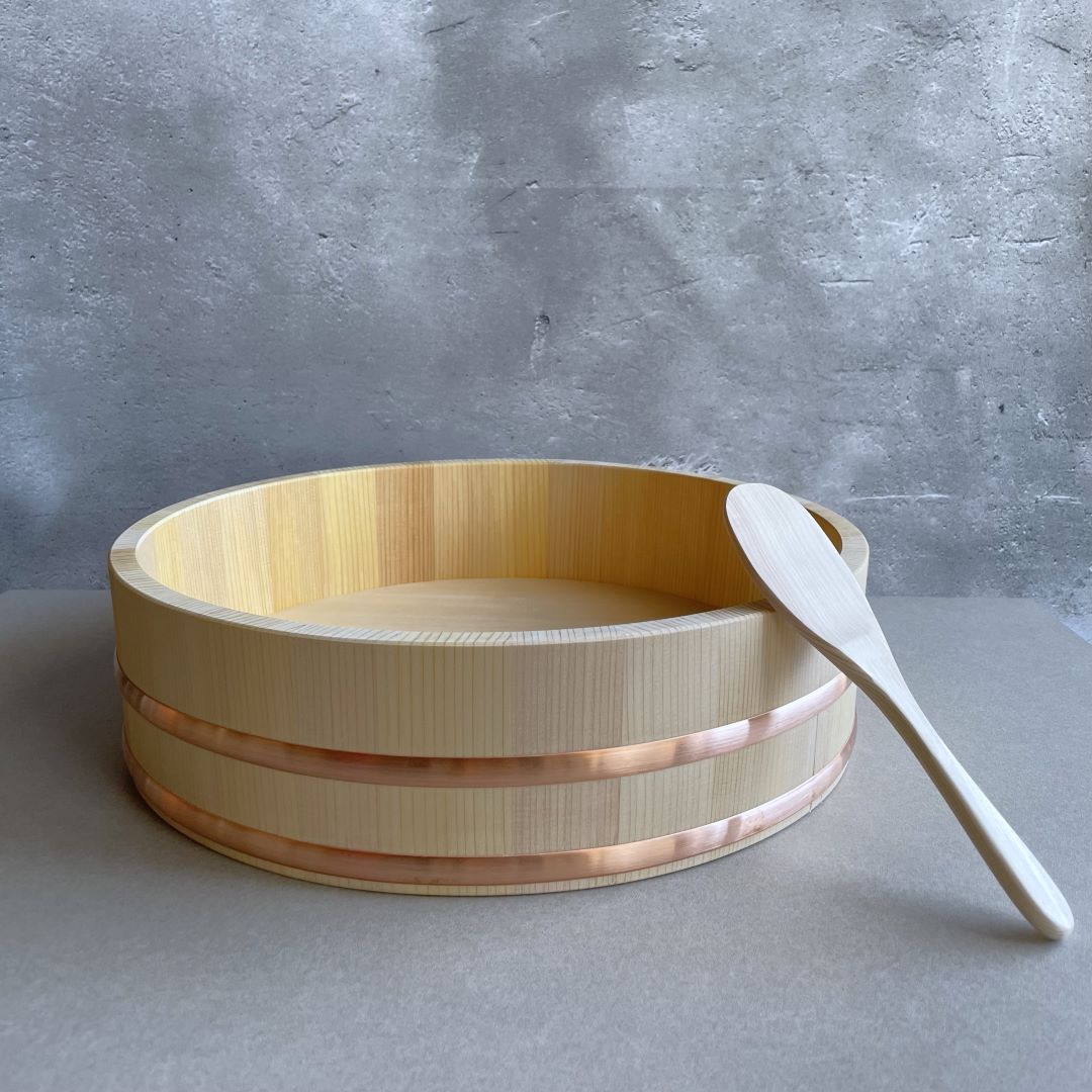 A Sawara cypress rice tub, traditionally used for preparing sushi rice, accompanied by a wooden paddle. The tub features copper bands and is designed for optimal rice cooling and flavor absorption.