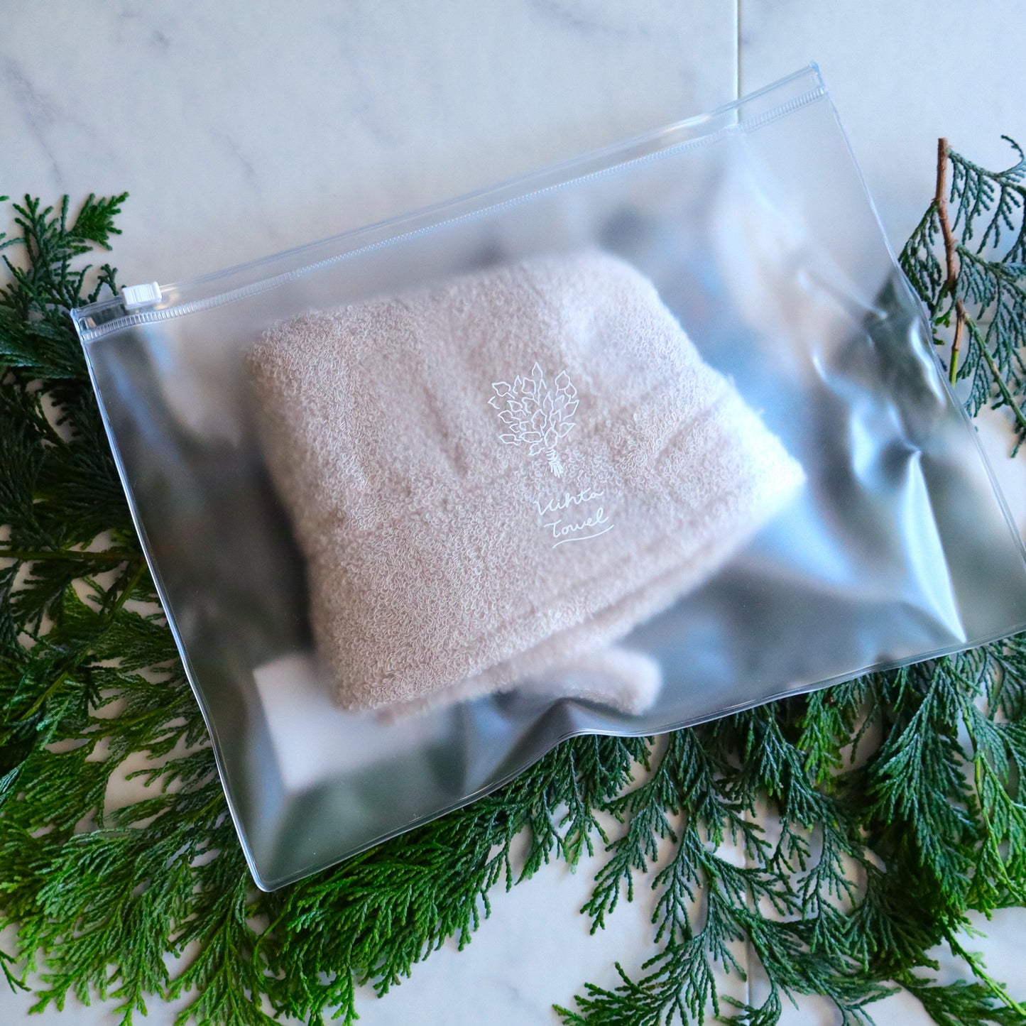 Beige sauna hat packed inside a translucent waterproof pouch, placed on fresh greenery.