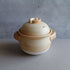 A beige and yellow Yamabuki-glazed ceramic donabe rice cooker with a lid, placed on a flat surface with a textured background.