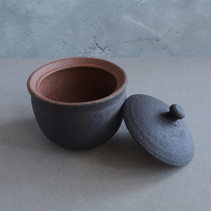 An angled close-up of a black ceramic salt cellar with its lid resting beside it, highlighting the smooth unglazed interior and the textured exterior with a contrasting brown rim.