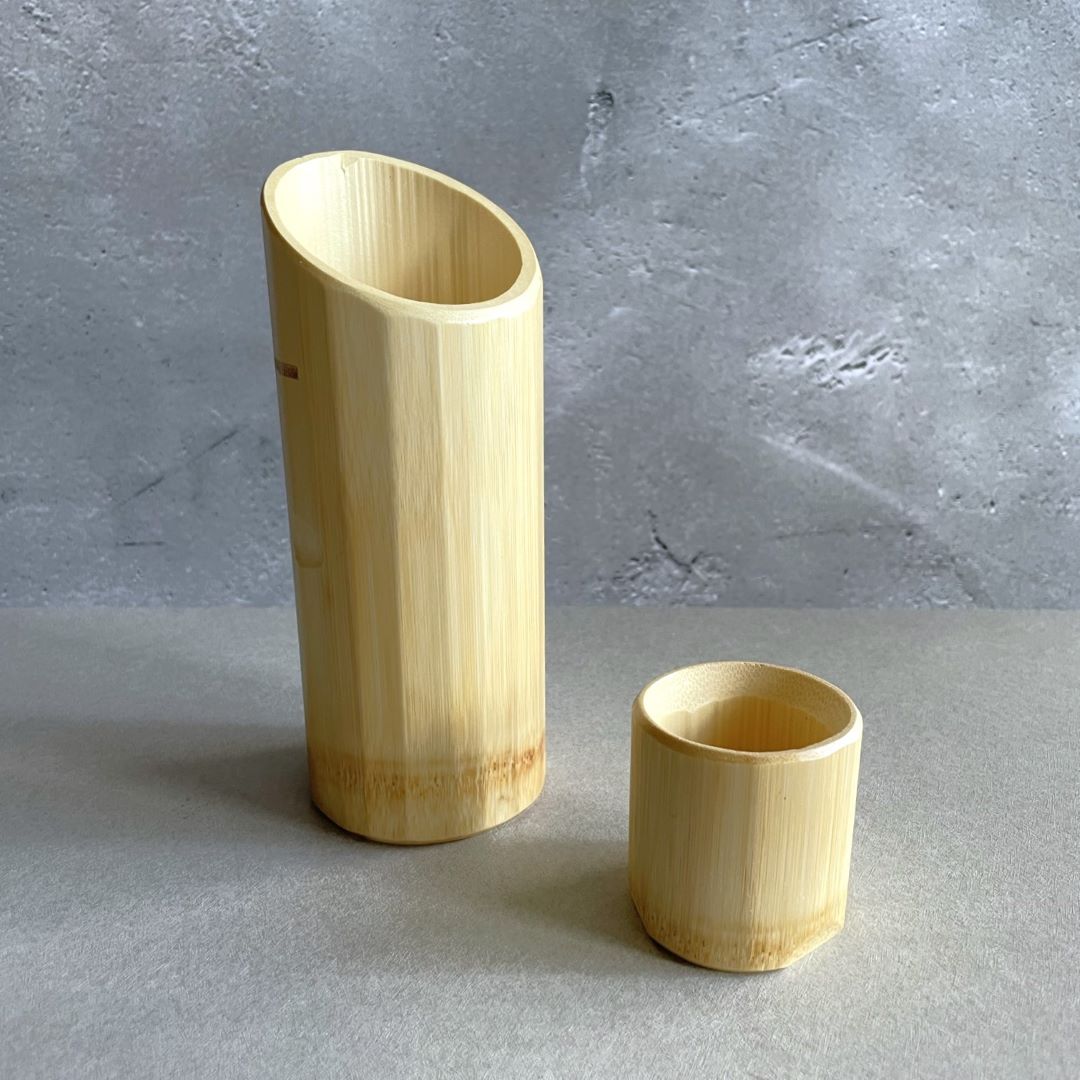 A tall bamboo vase and a shorter bamboo cup placed on a grey surface against a textured grey wall. The vase is positioned on the left with its height approximately twice that of the cup on the right. Both items display the natural vertical striations of bamboo and have a polished finish, with the vase showing a darker gradient at the bottom.