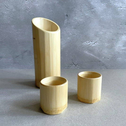Three bamboo containers of varying heights placed on a grey surface against a textured grey wall. The tallest container stands on the left and appears to be a vase, while the two shorter ones could be cups or holders, placed to the right with a small gap between them. The natural bamboo material shows vertical grain patterns and a smooth finish.