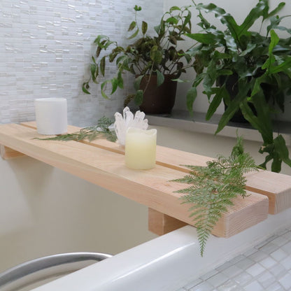 A Hinoki Cypress bathtub bench adorned with a white candle, a crystal cluster, and fresh greenery creates a spa-like atmosphere, laid across a bathtub with a background of soft-focus green plants and white mosaic tiles.