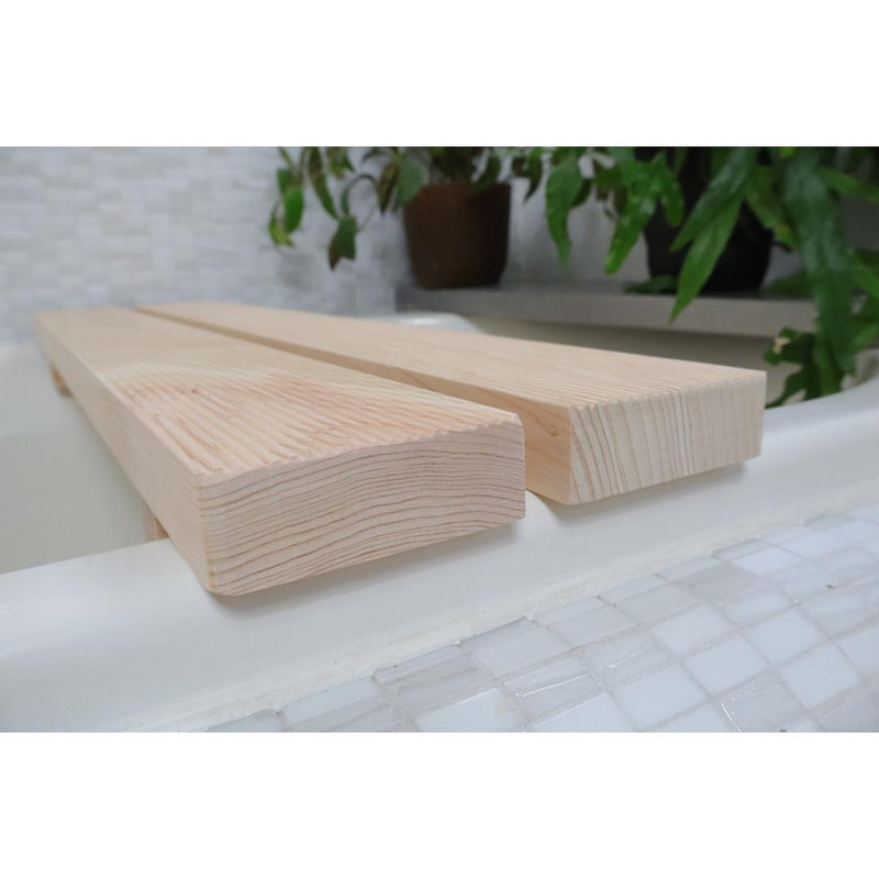 A minimalist Hinoki Cypress bathtub bench with a natural finish rests on the edge of a white tub, its fine wood grain prominent against the tiled bathroom setting and a potted green plant in the blurred background.