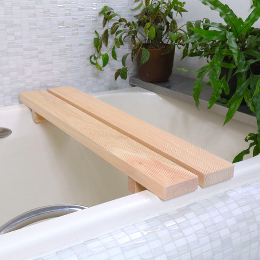 A light-hued Hinoki Cypress bathtub bench, also serving as a tub caddy, rests across a bathtub. At 27 inches long, it&#39;s perfectly sized for tubs between 20 to 23.6 inches wide, shown in a serene bathroom with a potted plant on a ledge.