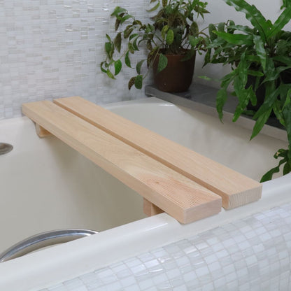  A minimalist Hinoki wood bath bench stretches across a white bathtub, juxtaposed against a mosaic tile backdrop with a lush potted plant, embodying a clean and tranquil spa setting.