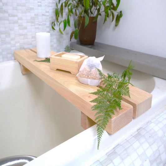 A light wooden bath bench spans across a bathtub, topped with a white candle, a wooden soap dish with soap, a bath aroma bag, and a sprig of fresh greenery, creating a peaceful, spa-like atmosphere in a bathroom with mosaic tiles and a potted plant in the background.