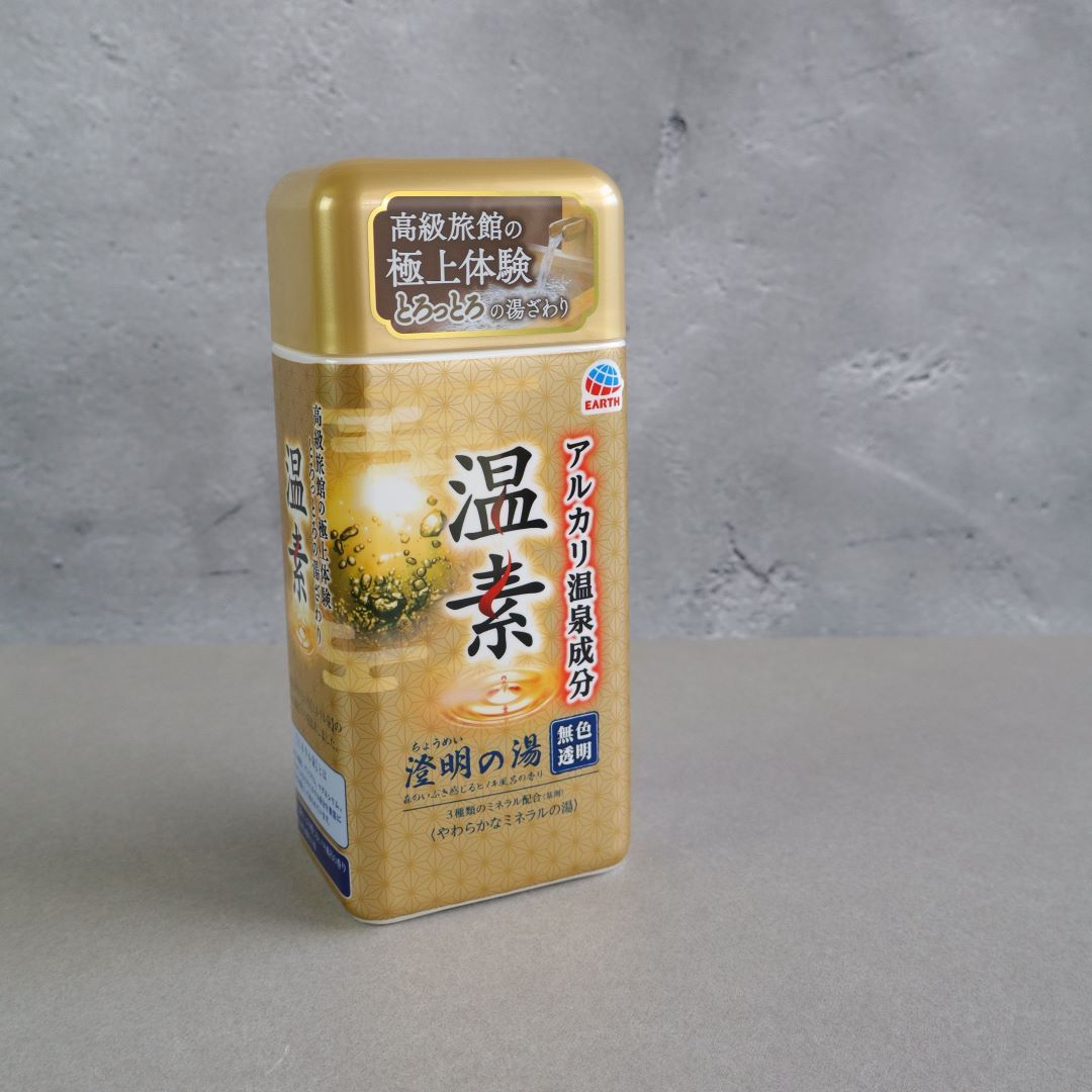 A container of Onso Alkaline Bath Salts standing upright against a neutral gray background. The packaging features a gold and brown design with Japanese text, branding from Earth, and an image of bubbling water, emphasizing its hot spring-inspired benefits. The product is designed to recreate the relaxing and skin-smoothing effects of a Japanese onsen bath at home.