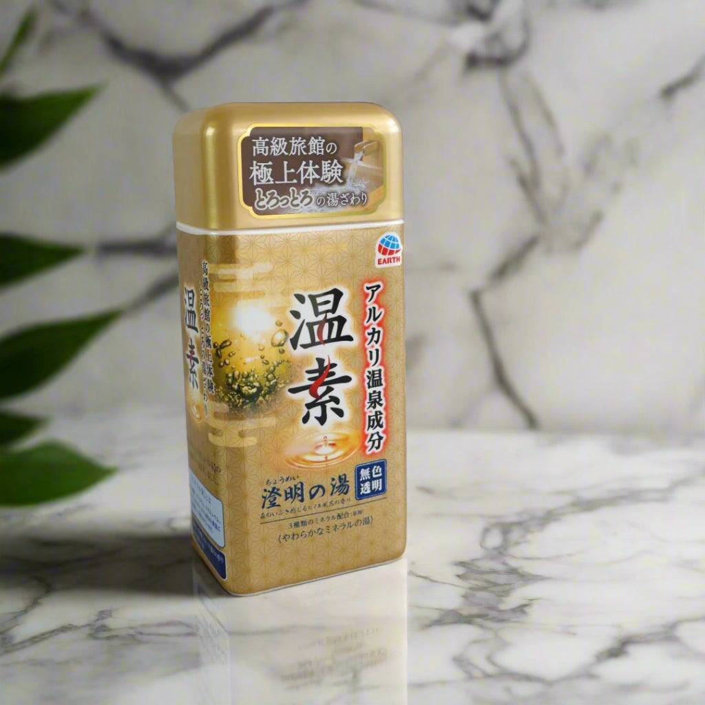 A container of Onso Alkaline Bath Salts standing upright against a neutral gray background. The packaging features a gold and brown design with Japanese text, branding from Earth, and an image of bubbling water, emphasizing its hot spring-inspired benefits. The product is designed to recreate the relaxing and skin-smoothing effects of a Japanese onsen bath at home.