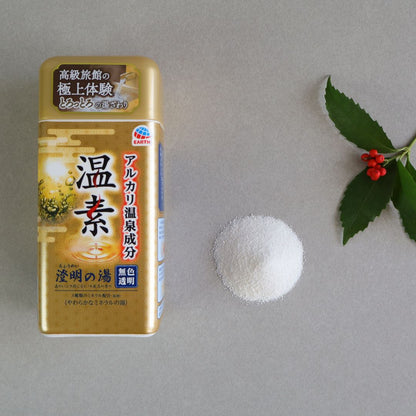 A container of Onso Alkaline Bath Salts with a gold and brown design, featuring Japanese text and branding from Earth. Next to the container is a small pile of fine white bath powder, and a sprig of green leaves with red berries is placed on the right. The background is a neutral gray surface, creating a clean and minimalist presentation.