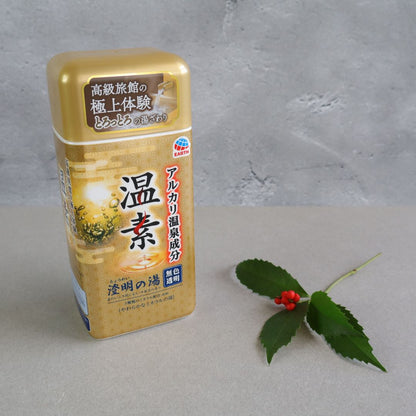 A container of Onso Alkaline Bath Salts standing on a neutral surface, with a sprig of green leaves and red berries beside it. The packaging features a gold and brown design with Japanese text, highlighting its hot spring-inspired benefits. The product aims to recreate the relaxing and skin-smoothing effects of a Japanese onsen bath at home. The background is a soft gray, complementing the product’s natural aesthetic.