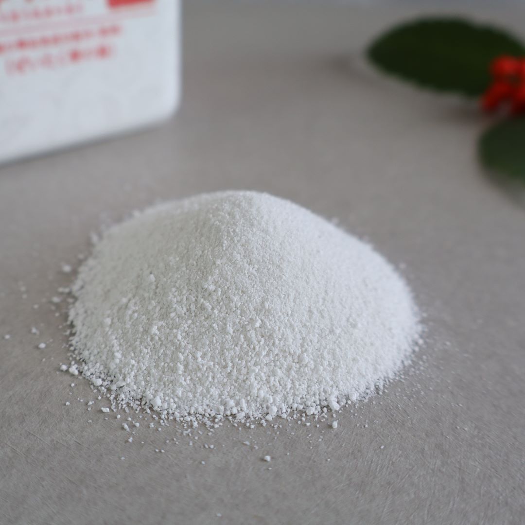 A close-up of a small pile of fine white bath salts, highlighting their texture. Partially visible in the background is the Onso Shiraha no Yu bath salt container and green leaves with red berries, evoking a natural and relaxing spa experience.