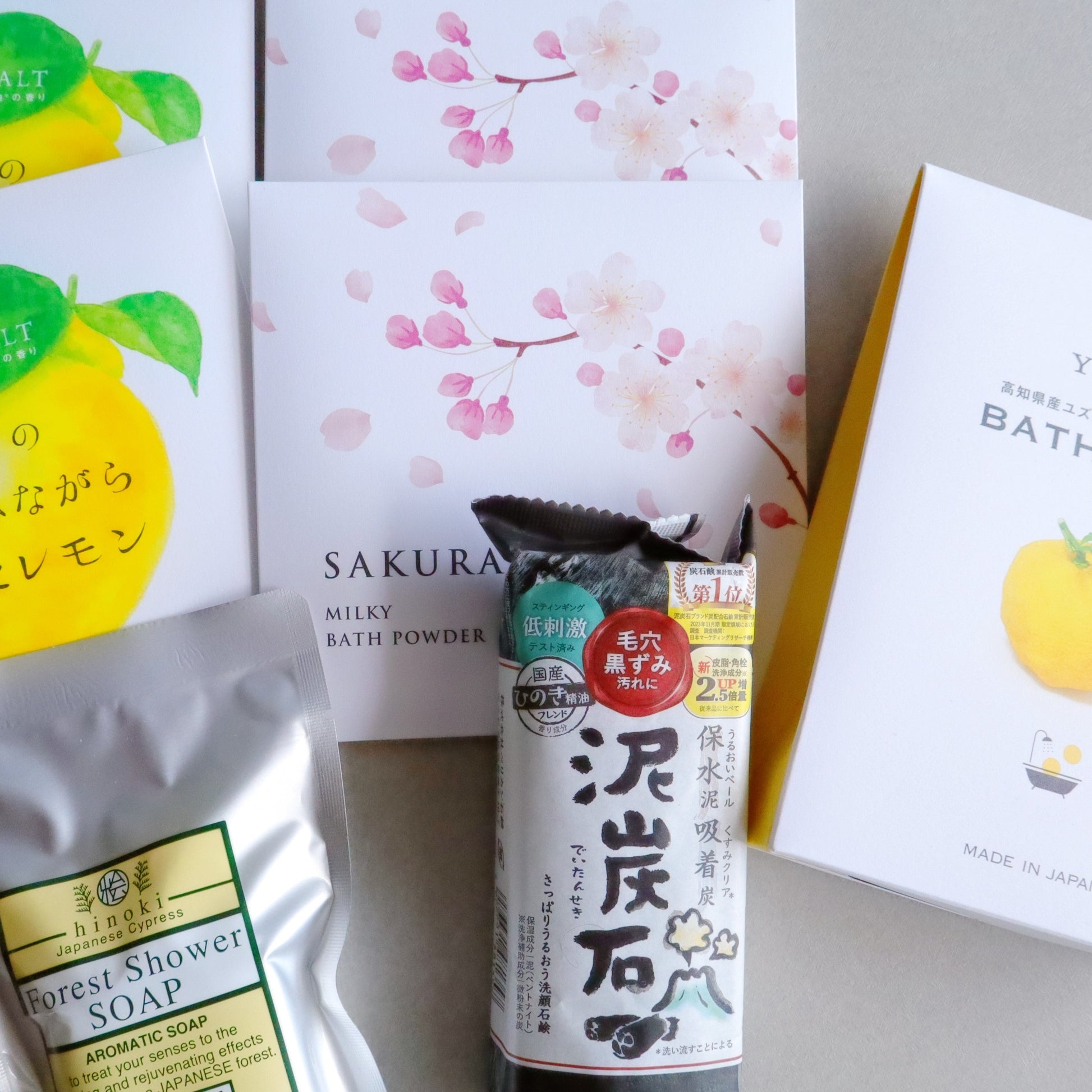 Japanese bath and skincare products arranged on a neutral surface, featuring Sakura Milky Bath Powder, Setouchi Lemon Bath Salt, Yuzu Bath Salt, a Hinoki Forest Shower Soap, and a Japanese charcoal soap bar. The packaging showcases elegant floral and citrus designs, emphasizing natural ingredients.