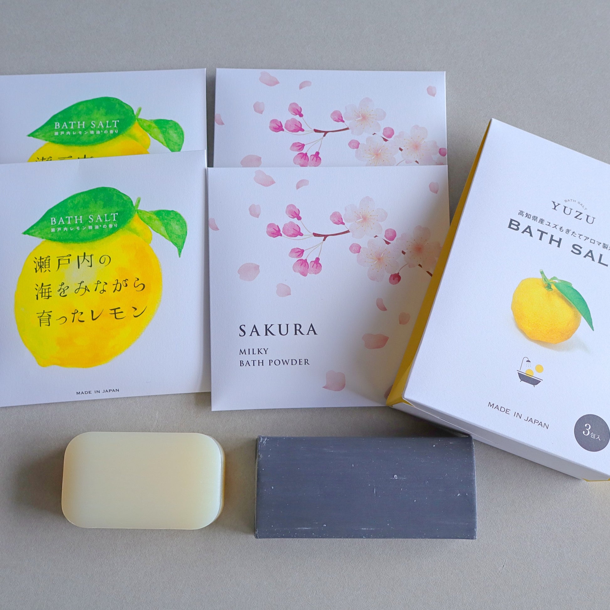 Japanese bath products displayed on a neutral background, including Sakura Milky Bath Powder, Setouchi Lemon Bath Salt, Yuzu Bath Salt, a white Hinoki soap bar, and a charcoal soap bar. The packaging features elegant floral and citrus-themed designs.