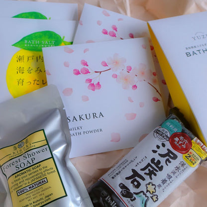 Japanese bath and body care products, including Sakura Milky Bath Powder, Yuzu Bath Salt, Setouchi Lemon Bath Salt, Forest Shower Soap with Hinoki, and a Japanese charcoal soap bar, arranged on soft wrapping paper.