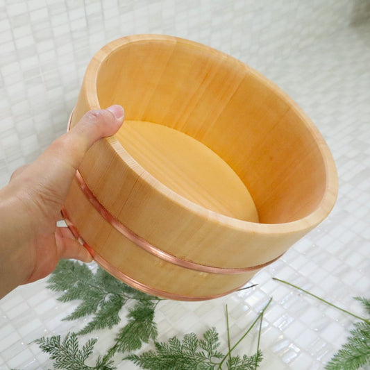 A hand holding a φ22cm / 8.66&quot; Japanese bath bucket made of Sawara cypress, showcasing its natural wood grain and smooth finish with a tile background and green ferns below.