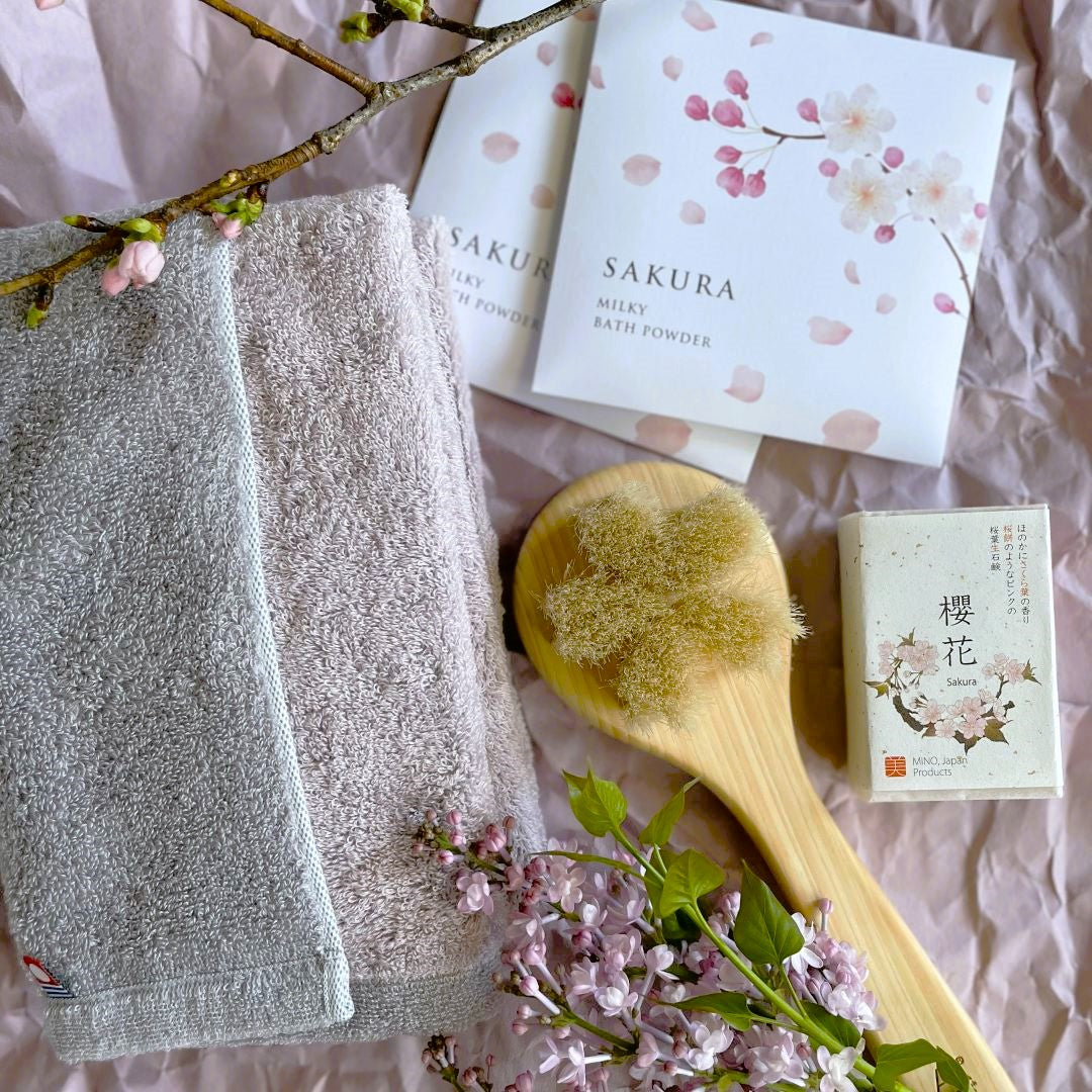 Japanese Sakura Bath Gift Set featuring an Imabari reversible towel, a natural wooden body brush, Sakura soap, and milky bath powder, elegantly arranged with cherry blossoms on soft pink wrapping paper.