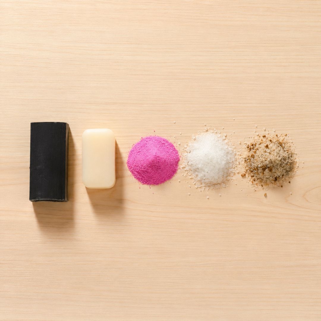 Four cosmetic or material samples arranged in a row on a light wood surface: a black rectangular block, a pale cream rectangular block, a bright pink powder pile, a white powder pile, and a brownish-beige textured powder pile.