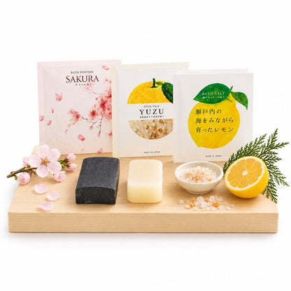 Three packaged bath salts are arranged on a light wood tray with corresponding decorative ingredients: a pink sakura pouch, a yellow yuzu pouch, and a green lemon pouch, alongside a dark soap bar, a pale soap bar, a small bowl of salt, and citrus and greenery on a white background.