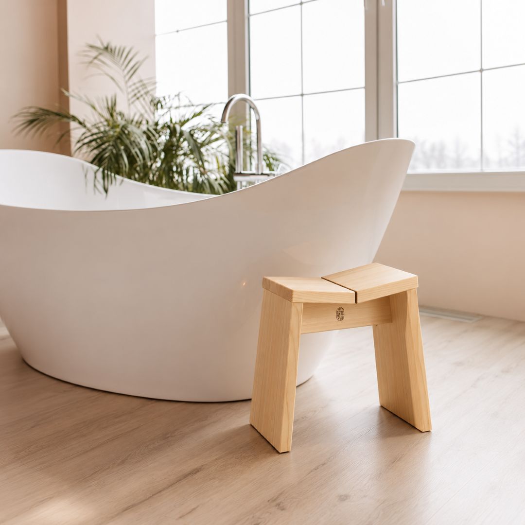 A small light natural wood step stool stands beside a white freestanding bathtub in a bright bathroom with wood flooring and a window.