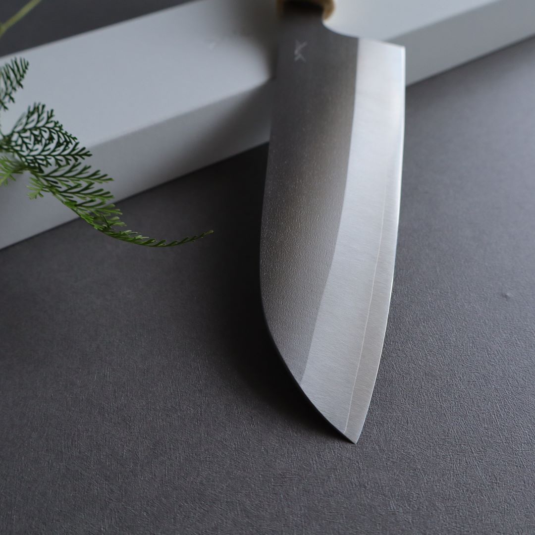 Close-up view of a Japanese knife with a stainless steel blade and a wooden handle. The blade is positioned on a gray surface, with a white rectangular object beneath the handle. A small sprig of green foliage is visible on the left side, adding a natural element to the composition.