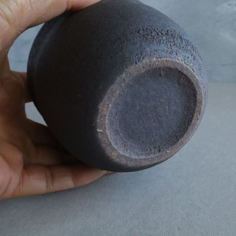 A hand holding a black ceramic salt cellar upside down to show the unglazed, textured bottom with a brown rim against a grey background.