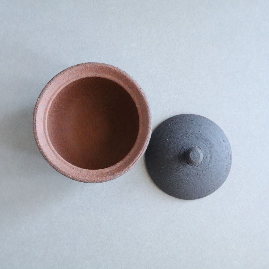 Top-down view of an open Shigaraki ceramic salt cellar with a dark exterior and a contrasting terracotta interior, alongside its round black lid, on a light grey background.